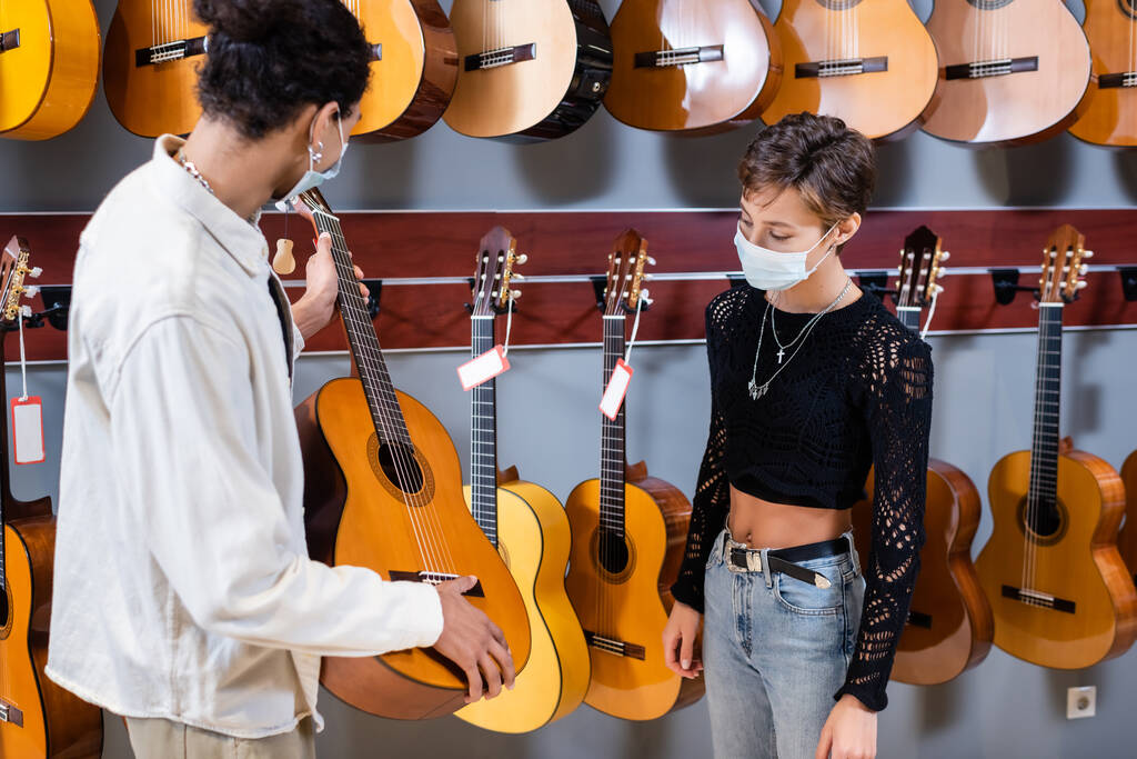 Müzik mağazasında tıbbi maskeli bir müşterinin yanında akustik gitar tutan Afrikalı Amerikalı bir satıcı.  - Fotoğraf, Görsel