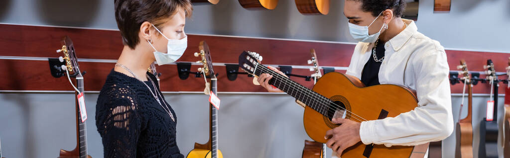 Customer in medical mask standing near african american seller playing acoustic guitar in store, banner  - Fotografie, Obrázek