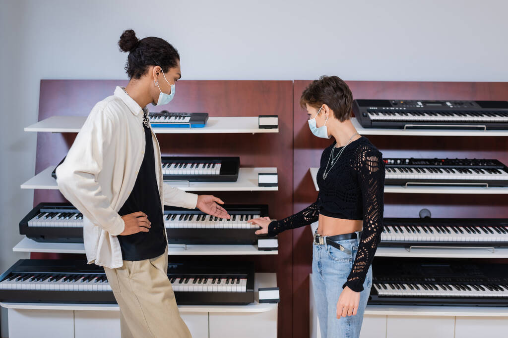 African american seller in medical mask pointing at synthesizer near customer in music store  - Foto, Imagen