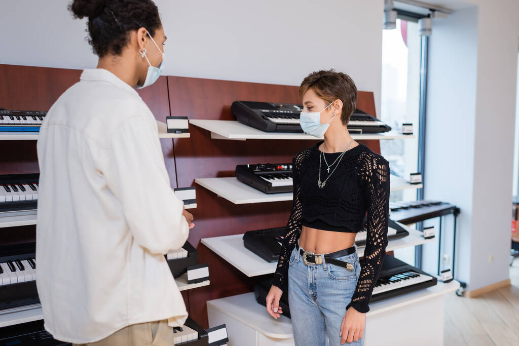 Klient w masce medycznej patrząc na syntezatory w pobliżu afrykańskiego amerykańskiego sprzedawcy w sklepie muzycznym  - Zdjęcie, obraz