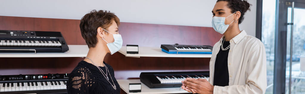 Afrikai amerikai eladó és ügyfél orvosi maszkok közelében álló szintetizátorok zeneboltban, banner  - Fotó, kép