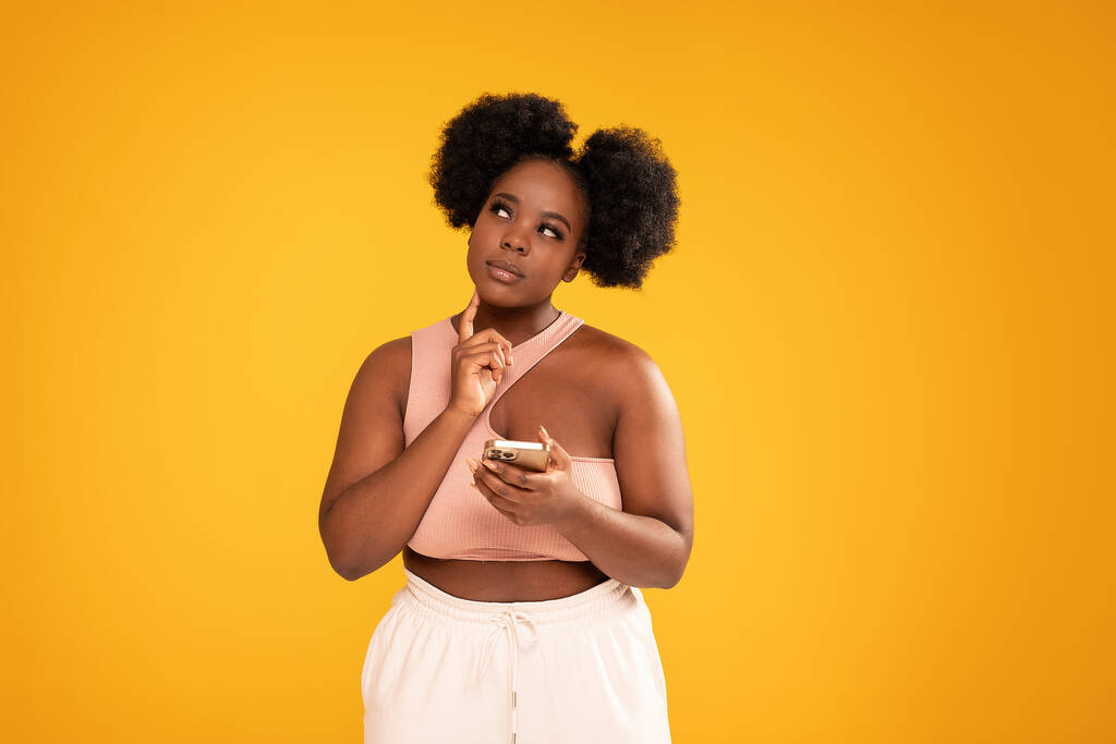Young afro woman holding mobile phone and thinking about buying something online. Sale concept. Studio shot. Yellow background - Foto, Imagen