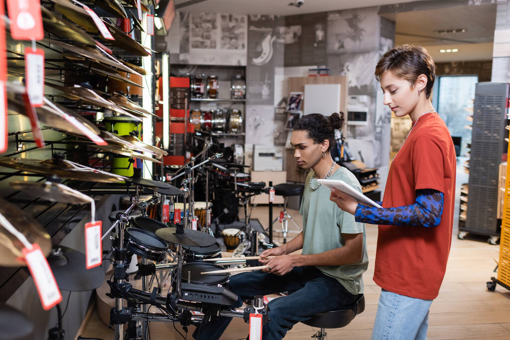 Вид сбоку продавца, держащего цифровой планшет рядом с покупателем, играющим в электрические барабаны в магазине  - Фото, изображение