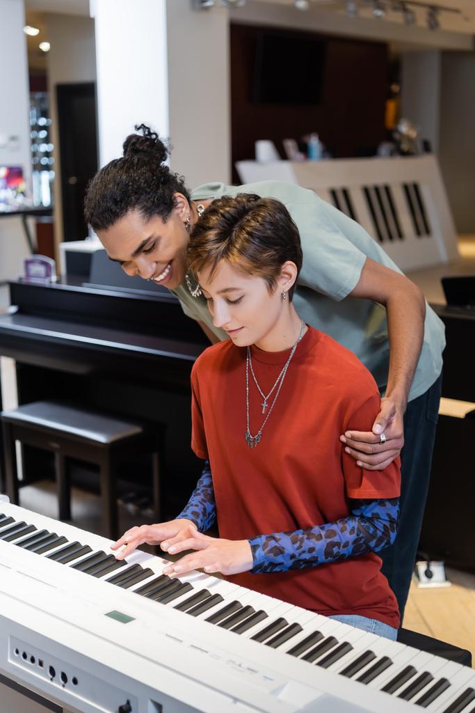 Cheerful african american man hugging girlfriend playing synthesizer in music shop  - Foto, Imagem