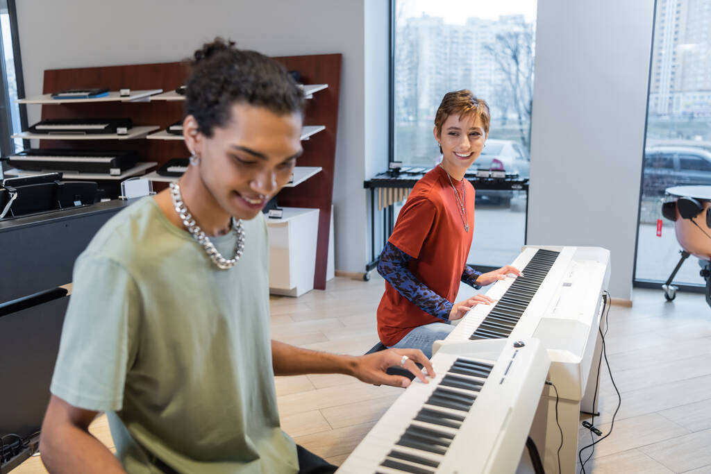 Cheerful woman playing synthesizer and looking at blurred african american boyfriend in music shop  - Foto, Imagen