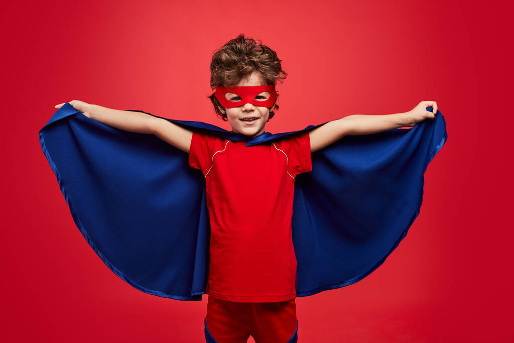Glad little boy in superhero costume looking at camera with smile and spreading cloak while playing against red background - Foto, Bild