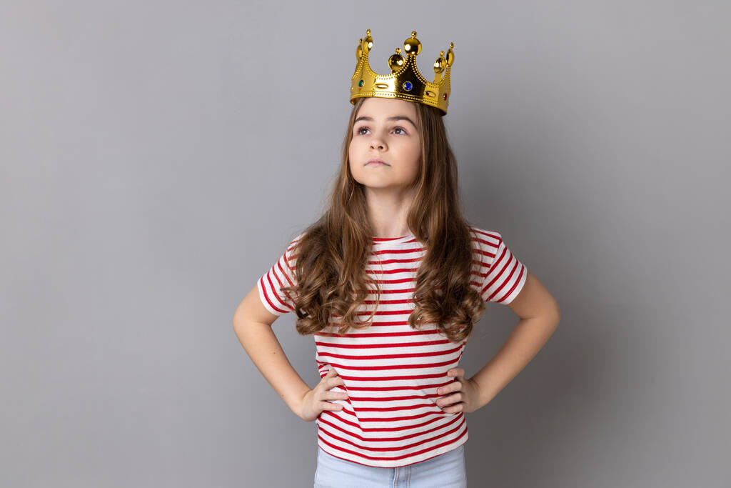 Portrait of confident little girl princess wearing striped T-shirt keeping hands on hips, self-motivation and dreams to be best. Indoor studio shot isolated on gray background. - Φωτογραφία, εικόνα