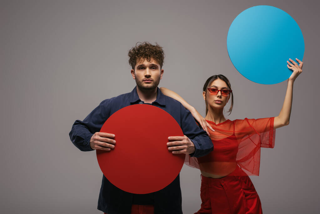 stylish models in trendy blue and red outfits holding round shape carton isolated on grey - Photo, Image