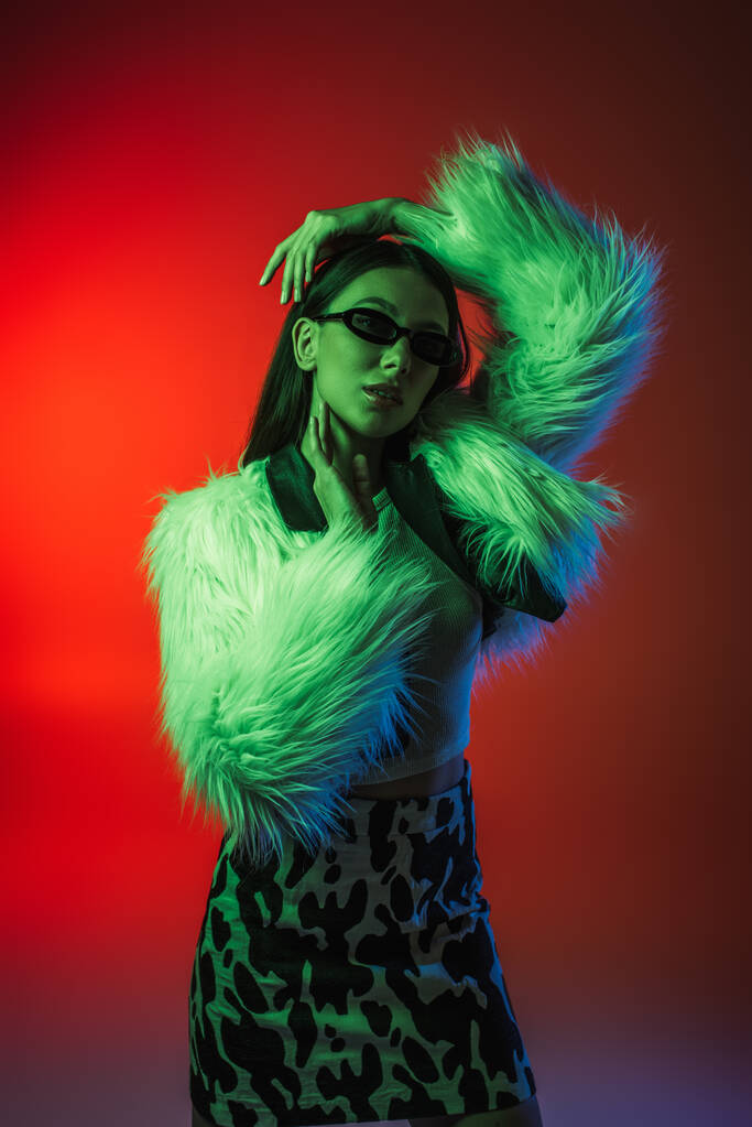 stylish young woman in faux fur jacket and sunglasses posing on red - 写真・画像