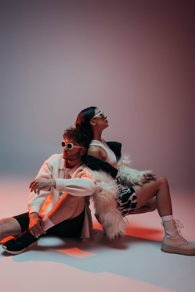 stylish man in sunglasses and woman in faux fur jacket sitting while posing on pink and grey - Photo, Image