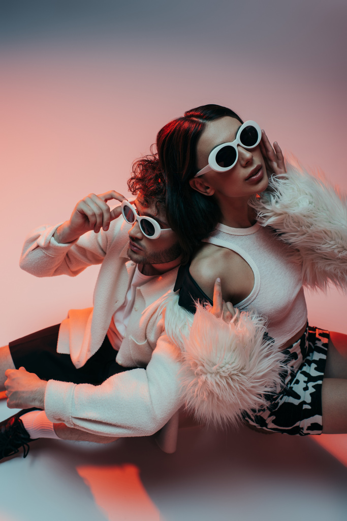 high angle view of stylish man in sunglasses leaning on woman in faux fur jacket on pink and grey - Zdjęcie, obraz