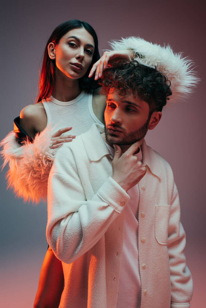 young woman in faux fur jacket gesturing near thoughtful man posing on grey - Photo, image