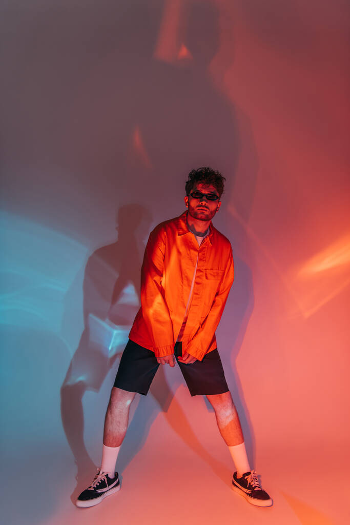 volledige lengte van krullende en stijlvolle man in een zonnebril poseren in de studio met kleurrijk licht - Foto, afbeelding