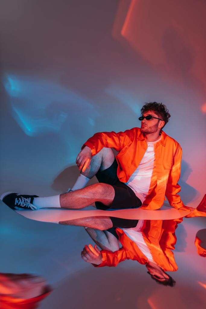 pełna długość kręcone i stylowy mężczyzna w okularach przeciwsłonecznych pozowanie podczas siedzenia w studio z kolorowym światłem - Zdjęcie, obraz