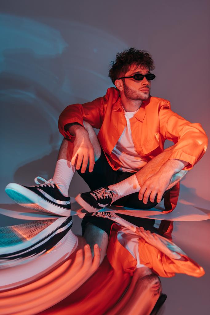täyspitkä trendikäs mies aurinkolasit poseeraa istuessaan studiossa värikäs valo - Valokuva, kuva