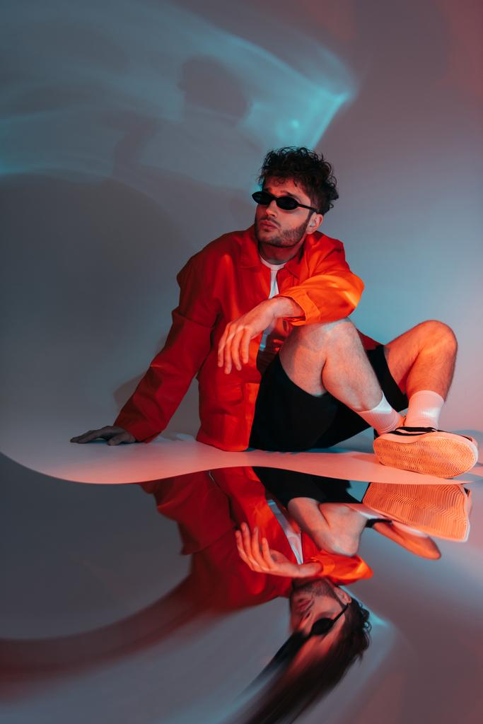 full length of curly man in sunglasses posing while sitting on grey with colorful light - Foto, Imagen