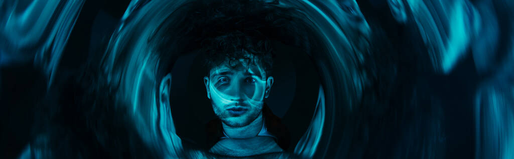 curly man looking at camera through futuristic neon blue circle on dark background, banner - Photo, image
