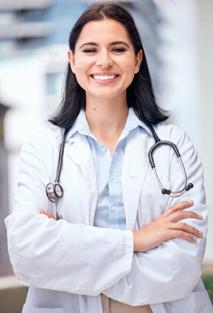 Confident young female doctor standing with her arms crossed outside a medical office. One caucasian woman in a white coat with stethoscope. Trusted practitioner caring for the health of patients. - Foto, Imagem