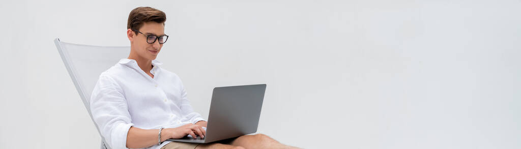 щасливий чоловік в окулярах використовує ноутбук у кріслі палуби ізольований на білому, банер
 - Фото, зображення