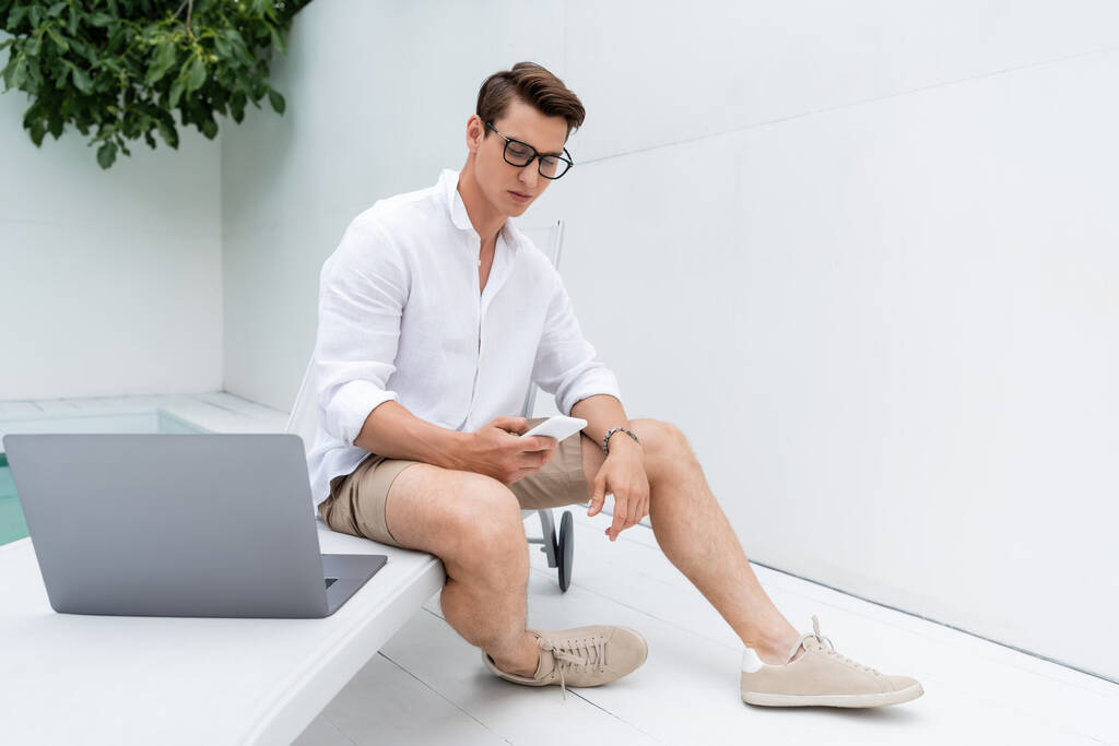 full length of man in sneakers sitting at poolside near laptop and messaging on mobile phone  - Foto, Imagen
