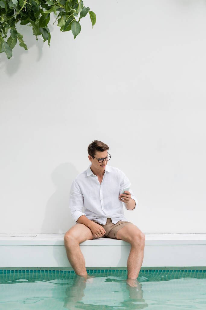 Lächelnder Mann sitzt am Pool an weißer Wand und schaut aufs Smartphone - Foto, Bild