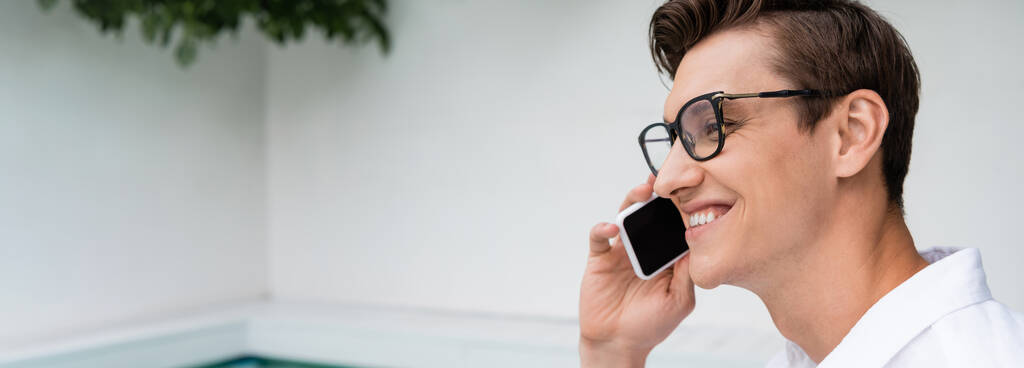 positive man in eyeglasses smiling while talking on smartphone outdoors, banner - Photo, image