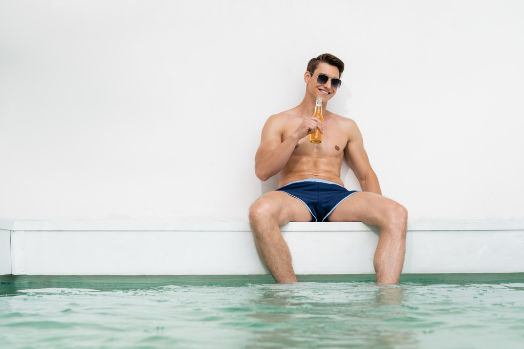 happy man in sunglasses and swimming trunks drinking beer near pool by white wall - Photo, image