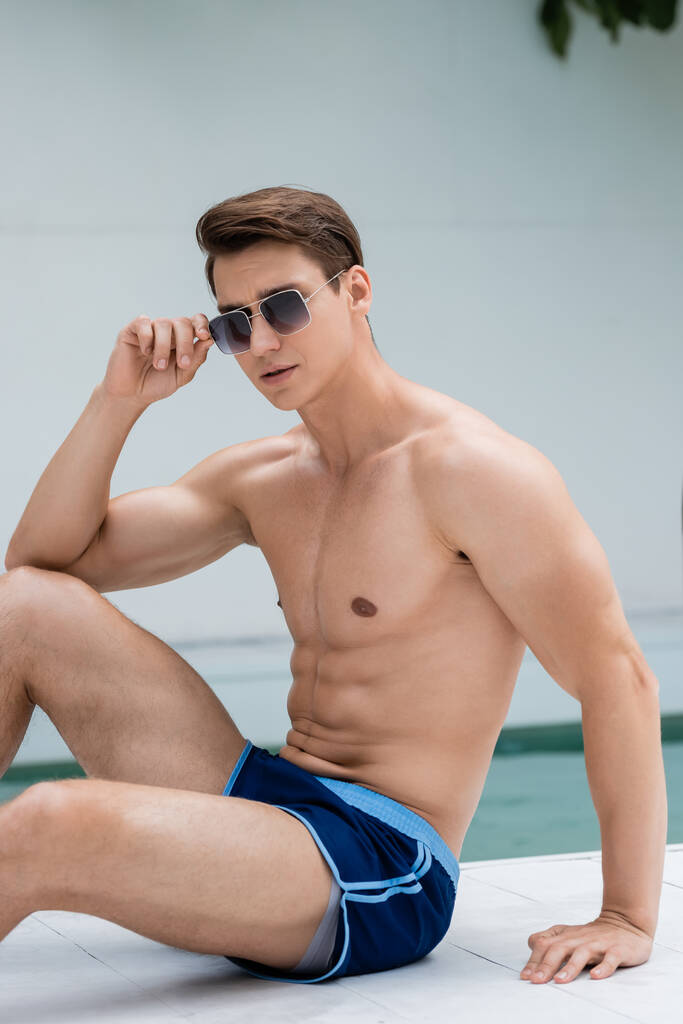 Мужчина без рубашки в спортивном теле регулирует солнцезащитные очки, сидя у бассейна - Фото, изображение