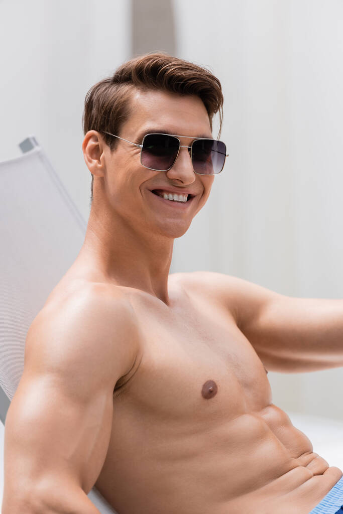 shirtloze man met een zonnebril glimlachend naar de camera terwijl hij in de ligstoel zit - Foto, afbeelding
