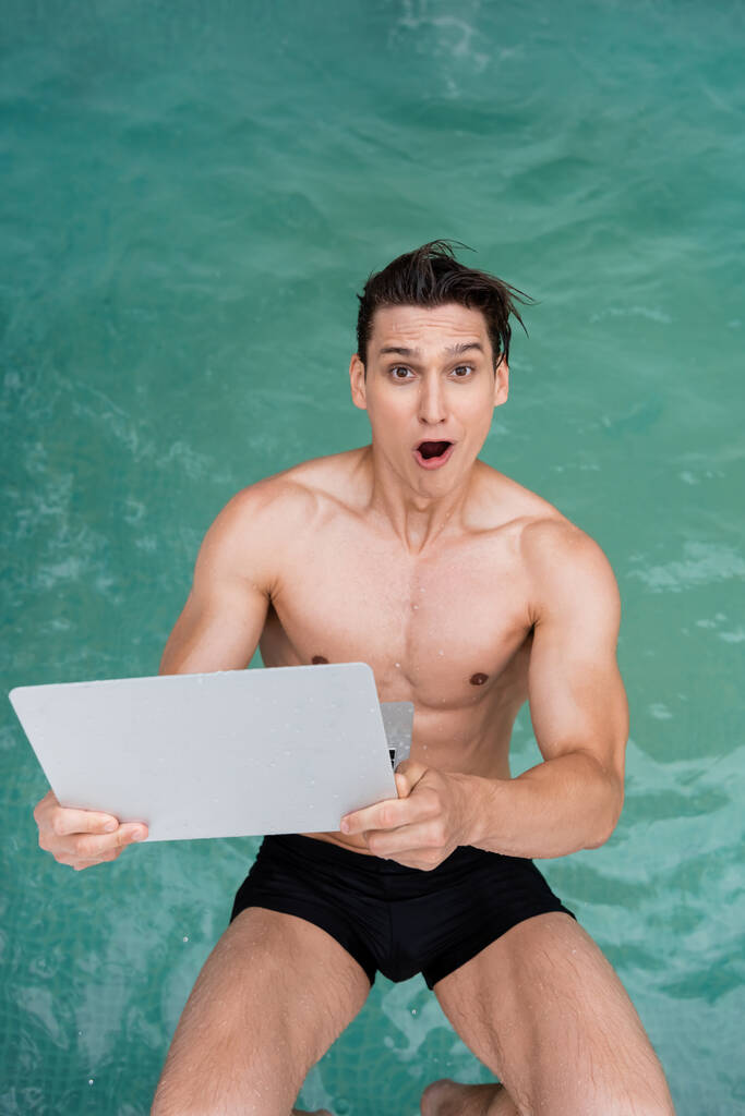 top view of astonished man with laptop looking at camera while falling in pool - Photo, image