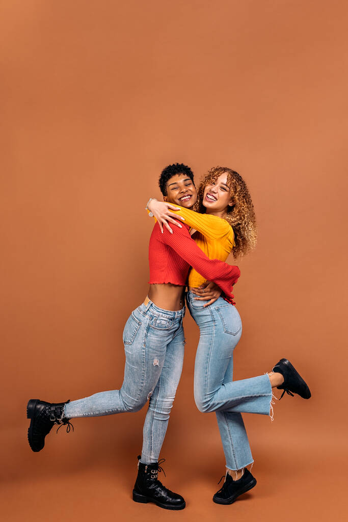 Stock photo of cheerful afro women hugging each other in studio shot against brown background. - 写真・画像