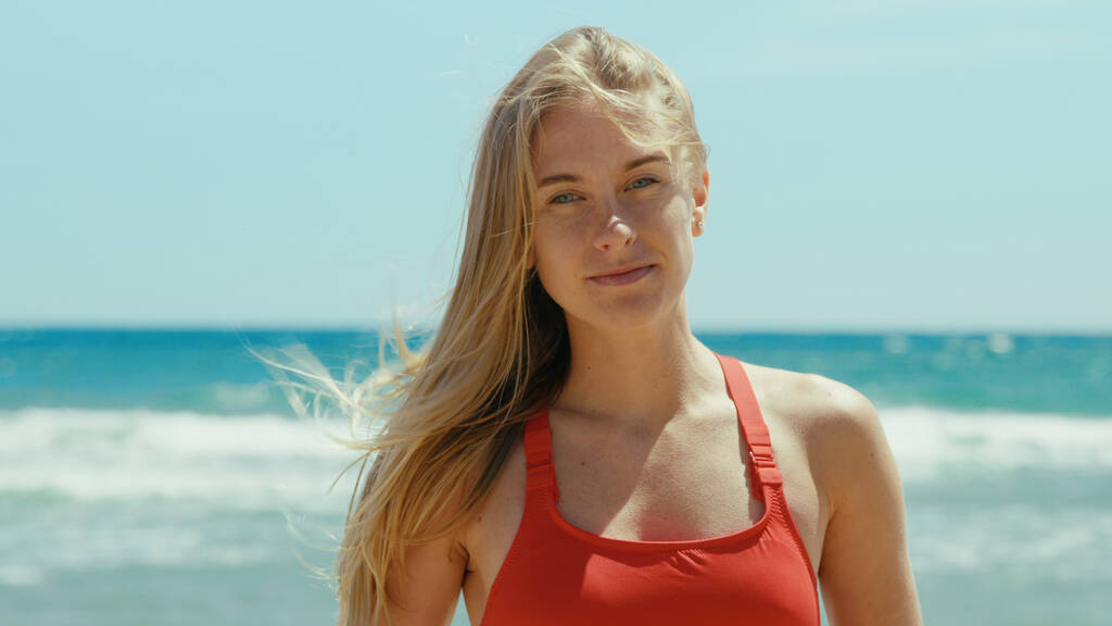 Retrato de una hermosa mujer joven en traje de baño rojo de pie en la playa soleada vacía, mirar a la cámara con confianza. Torre de vigilancia o guardia costera en segundo plano. Concepto de verano. Brisa cálida en el cabello - Foto, Imagen