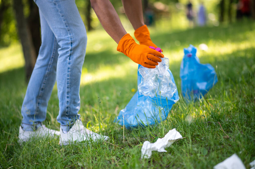 Care. Gloved hands of dark-skinned man putting plastic waste into bag on grass in park on sunny day without face - Zdjęcie, obraz