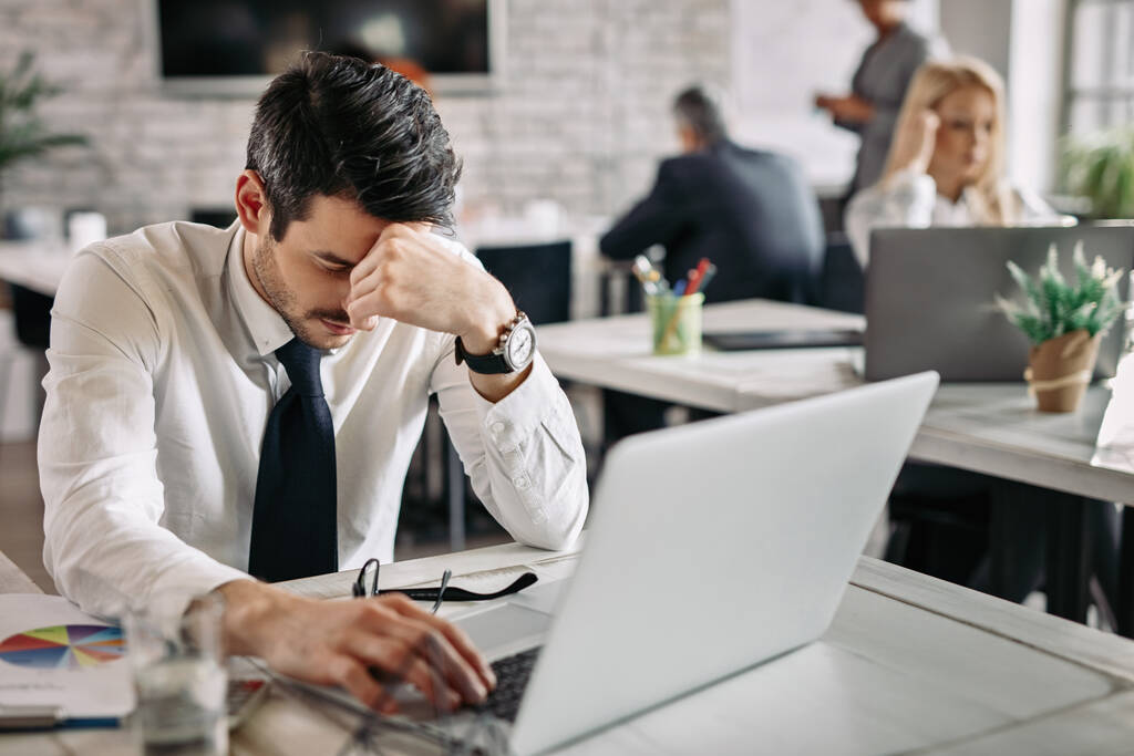 Tired businessman holding his head in pain while working on laptop and feeling exhausted from work. There are people in the background.  - Foto, immagini