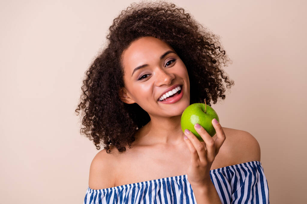 Foto di dolce bella ragazza vestita fuori spalle vestiti mangiare mela verde sorridente isolato colore beige sfondo. - Foto, immagini