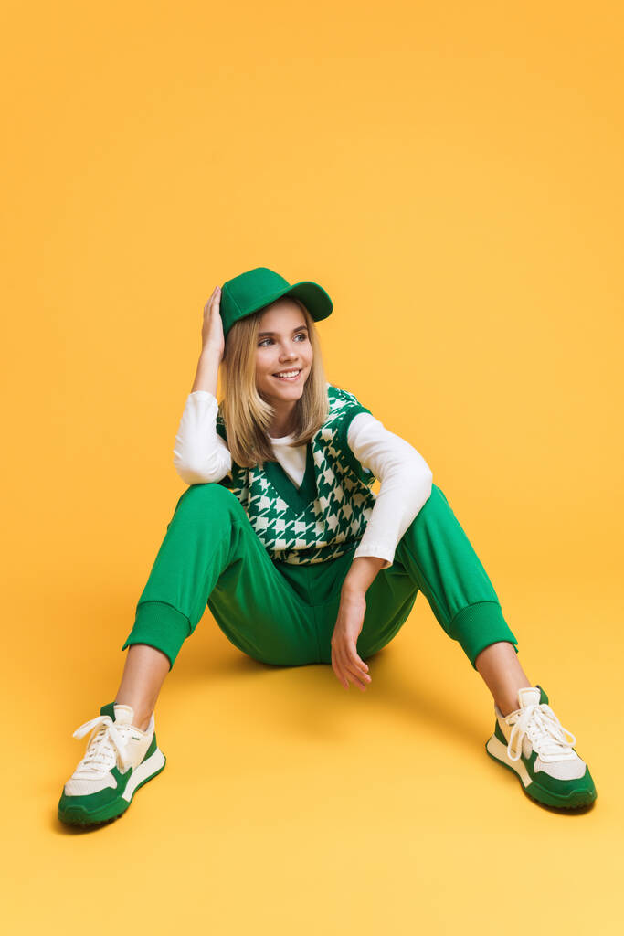White girl wearing vest smiling and sitting on floor isolated over yellow background - Photo, Image