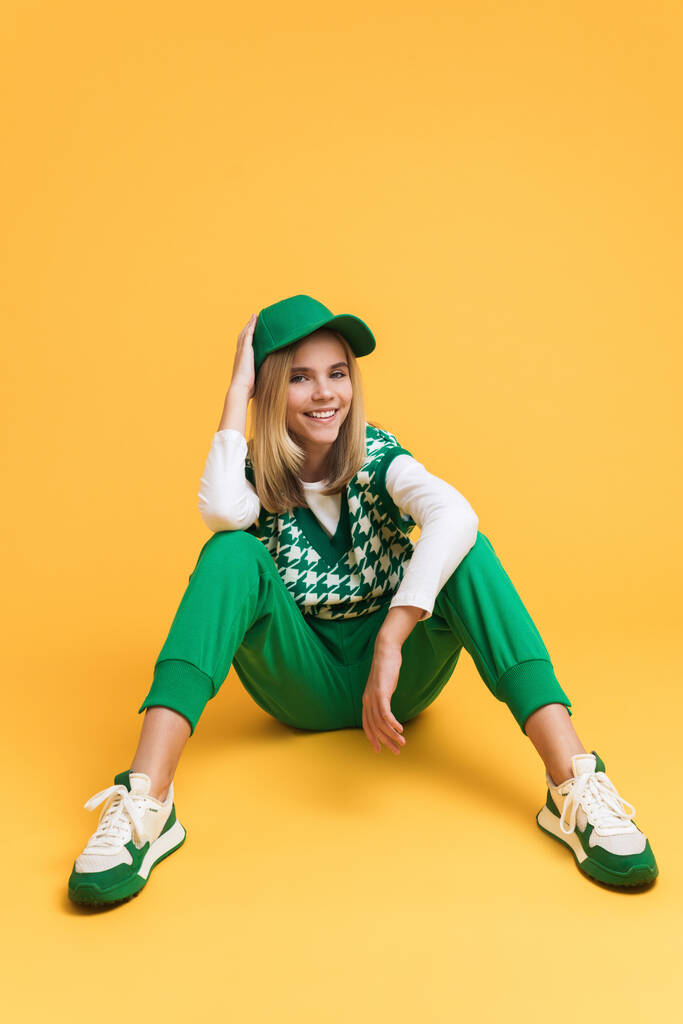White girl wearing vest smiling and sitting on floor isolated over yellow background - Φωτογραφία, εικόνα