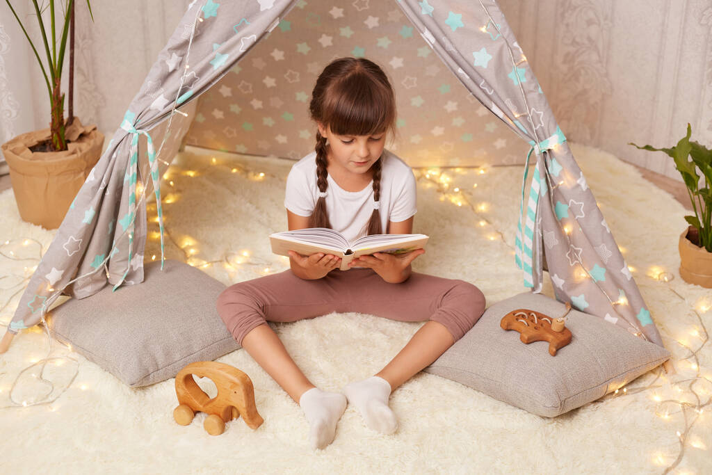 Horizontal shot of little girl wearing white t shirt reading book with pleasure and looking at pictures with interest, sitting in children's toy wigwam, child is at home. - Photo, Image