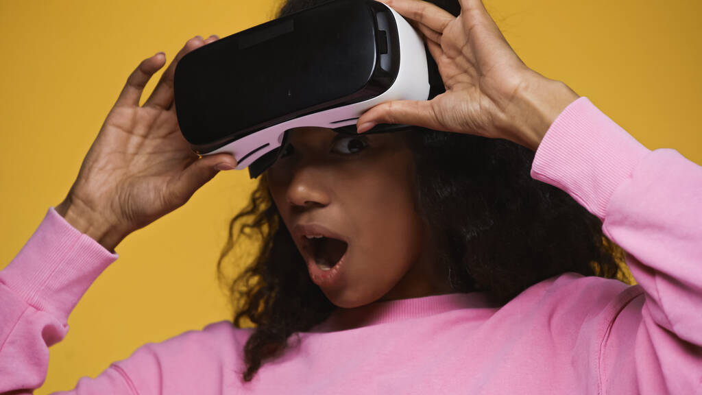 shocked african american woman in pink sweatshirt holding vr headset isolated on yellow - Foto, immagini