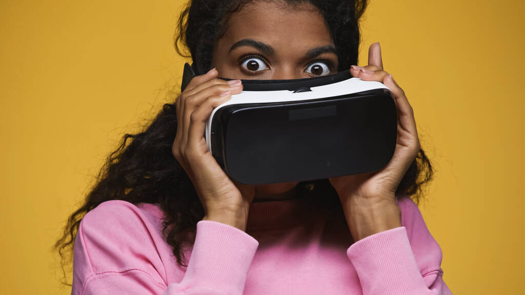 surprised african american woman in pink sweatshirt holding vr headset isolated on yellow  - Foto, afbeelding