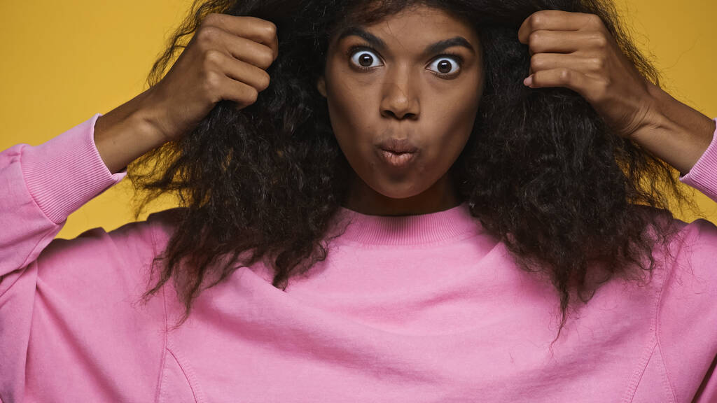 african american woman in pink sweatshirt pouting lips and adjusting curly hair isolated on yellow - Fotoğraf, Görsel