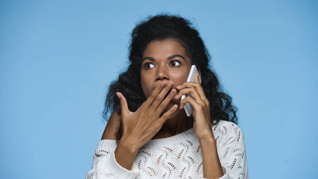 chocado afro-americano mulher em malha camisola cobrindo boca enquanto fala no smartphone isolado no azul - Foto, Imagem