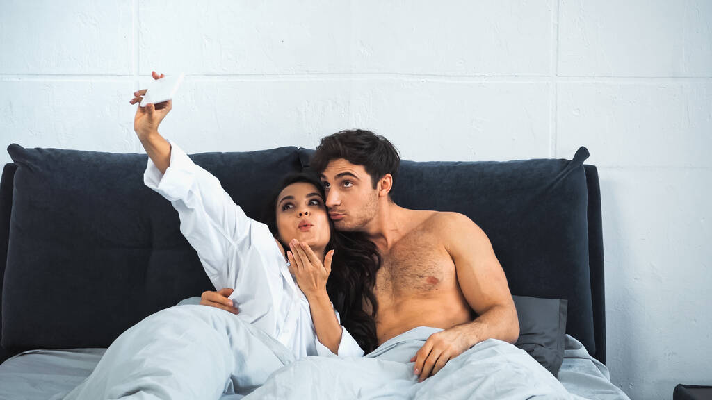 junge Frau schickt Luftkuss, während sie Selfie mit Mann ohne Hemd im Bett macht - Foto, Bild