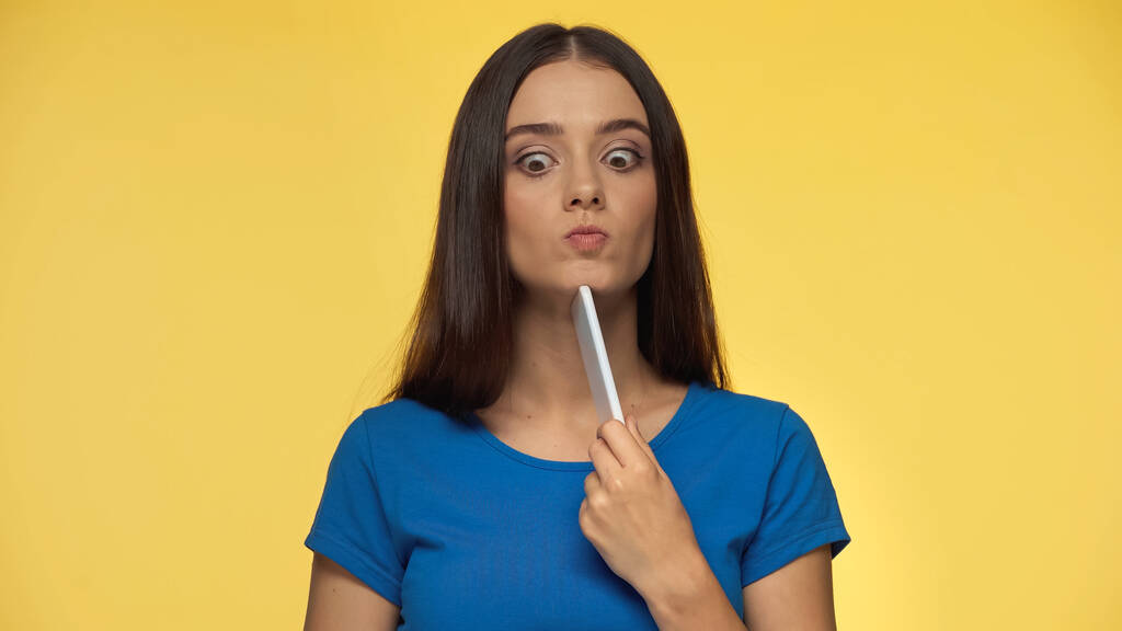 puzzled young woman in blue t-shirt pouting lips and holding smartphone isolated on yellow - Foto, afbeelding