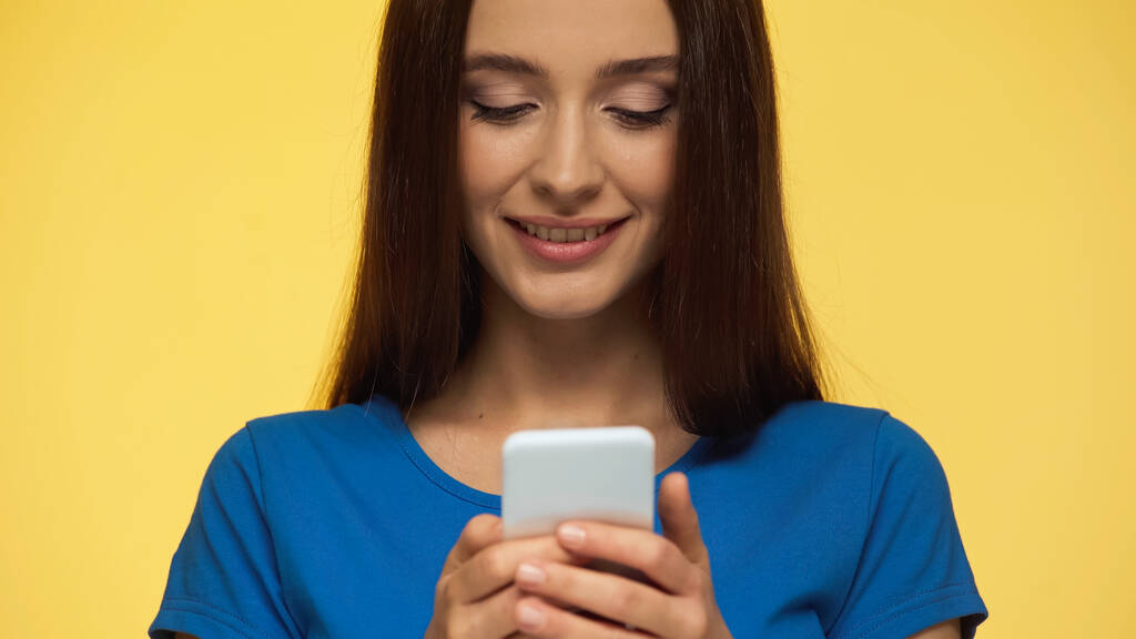 fiatal barna nő mosolyog, miközben csevegés mobiltelefon elszigetelt sárga - Fotó, kép