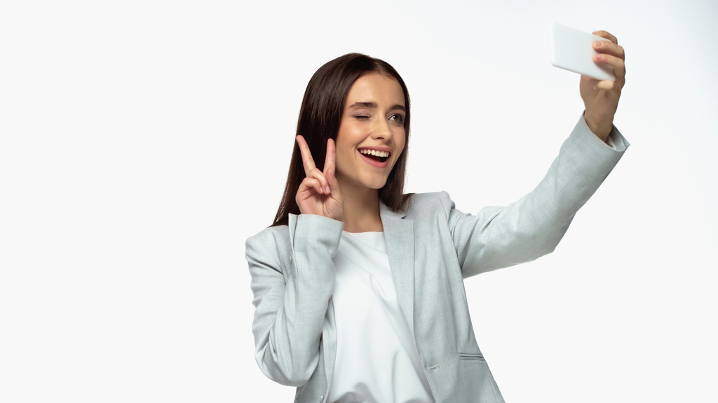 fröhliche Geschäftsfrau im grauen Blazer zeigt Friedenszeichen und macht Selfie isoliert auf Weiß - Foto, Bild