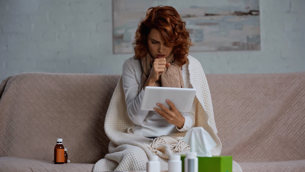 redhead sick woman using digital tablet and coughing near medication while sitting on couch - Photo, image