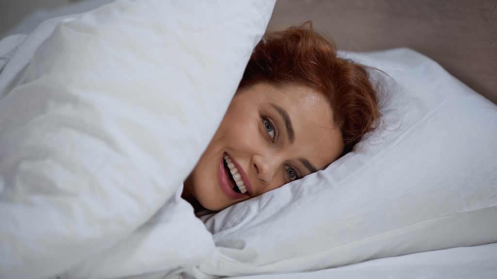 cheerful and redhead woman lying on pillow under white blanket - Photo, Image