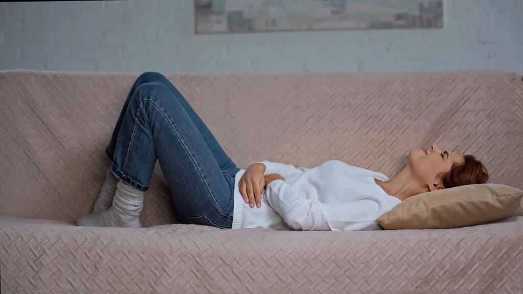Genç bir kadın kanepeye uzanıyor ve karın ağrısı çekiyor. - Fotoğraf, Görsel