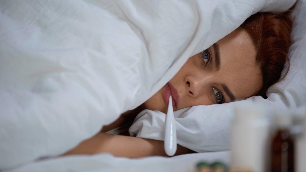 хвора жінка вимірює температуру з електронним термометром і лежить на ліжку
 - Фото, зображення